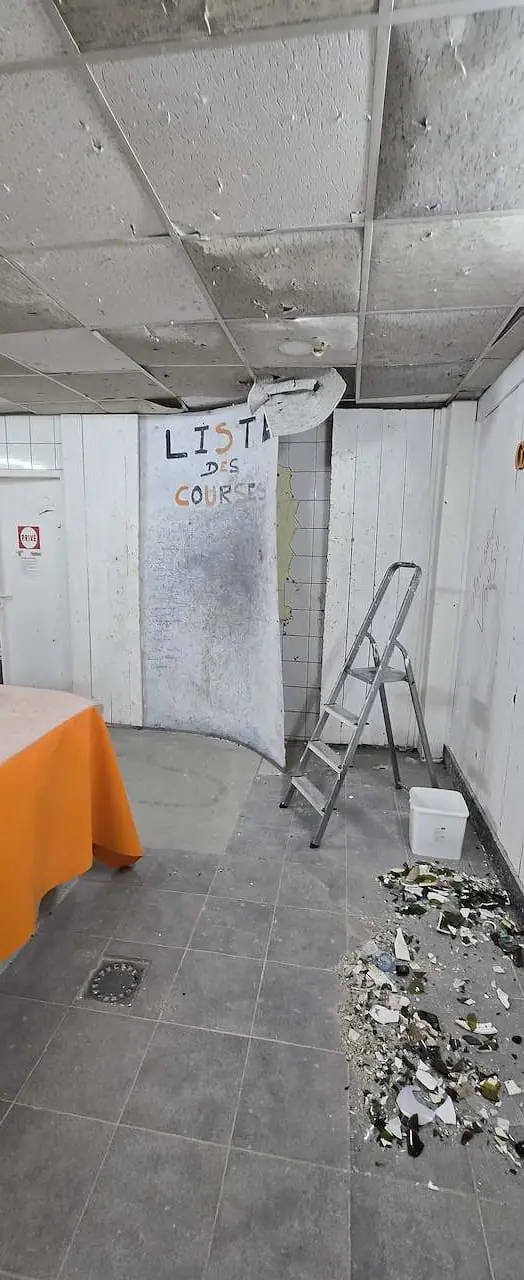 Débris de verre et de vaisselle au sol d'une salle de L'Atelier KS, près d'un escabeau et d'un panneau « Liste des courses »