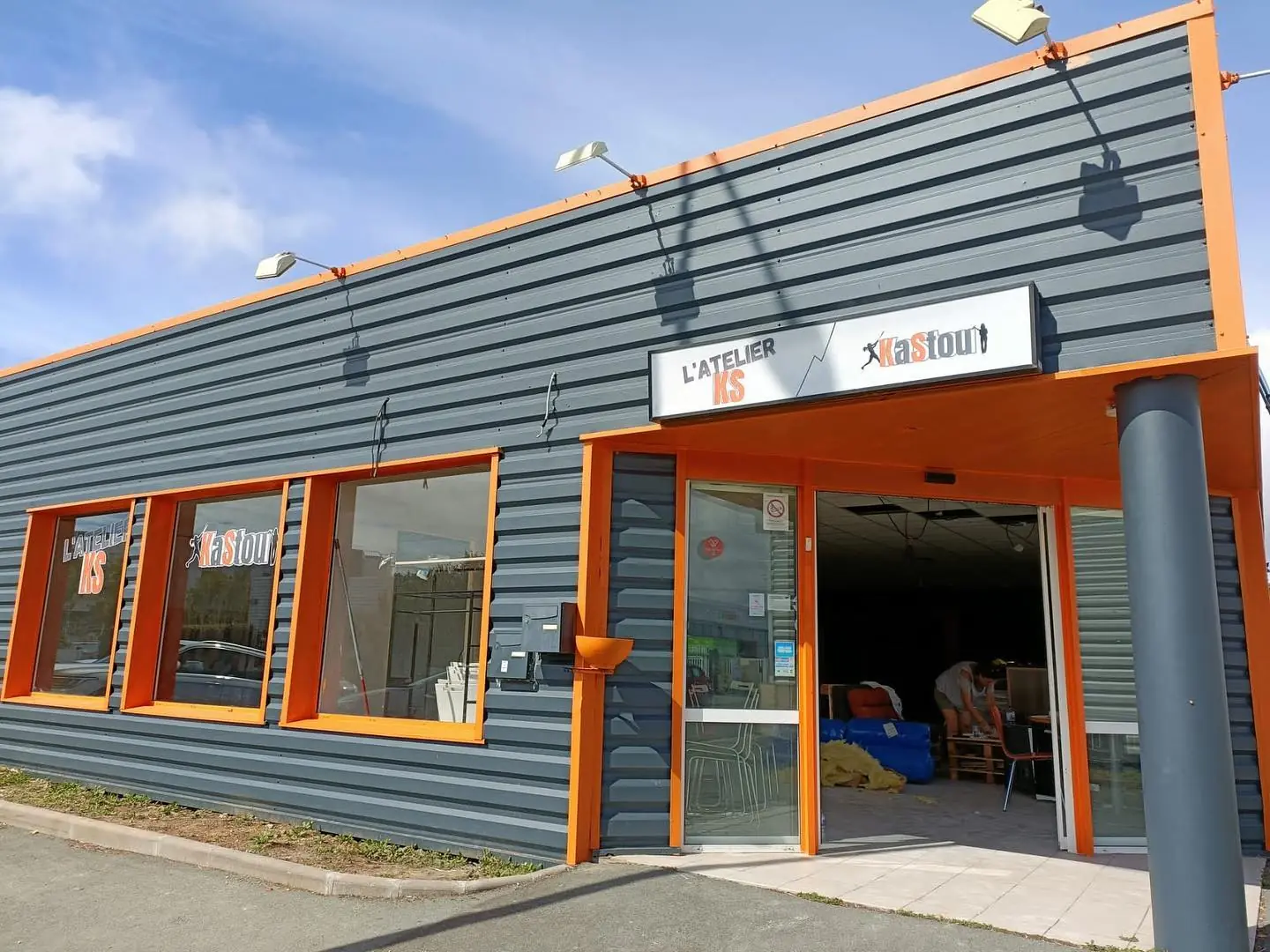 Façade grise et orange de L'Atelier KS à La Rochelle, porte ouverte sur l'accueil où des meubles en palettes sont en cours de montage