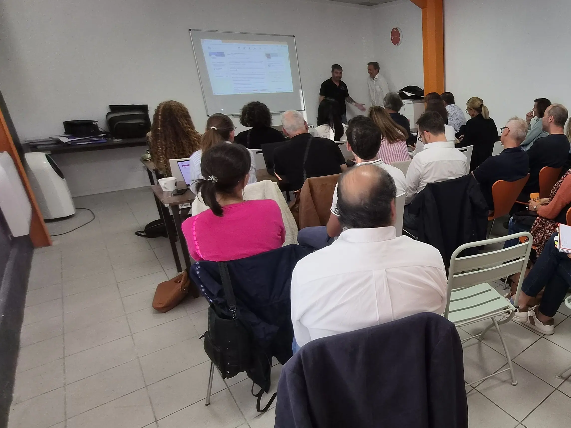 Une réunion dans la salle de L'Atelier KS : une vingtaine de participants assis de dos, face à deux intervenants et à un écran projeté.