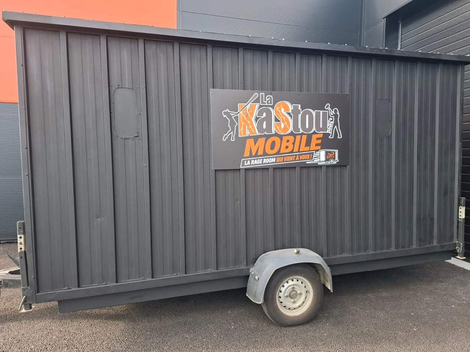 La Kastou Mobile, la rage room mobile de L'Atelier KS : une remorque noire à un essieu, habillée de tôle ondulée, garée sur un parking devant un bâtiment gris et orange. Sur son flanc, un panneau : « La Kastou Mobile, la rage room qui vient à vous ! »