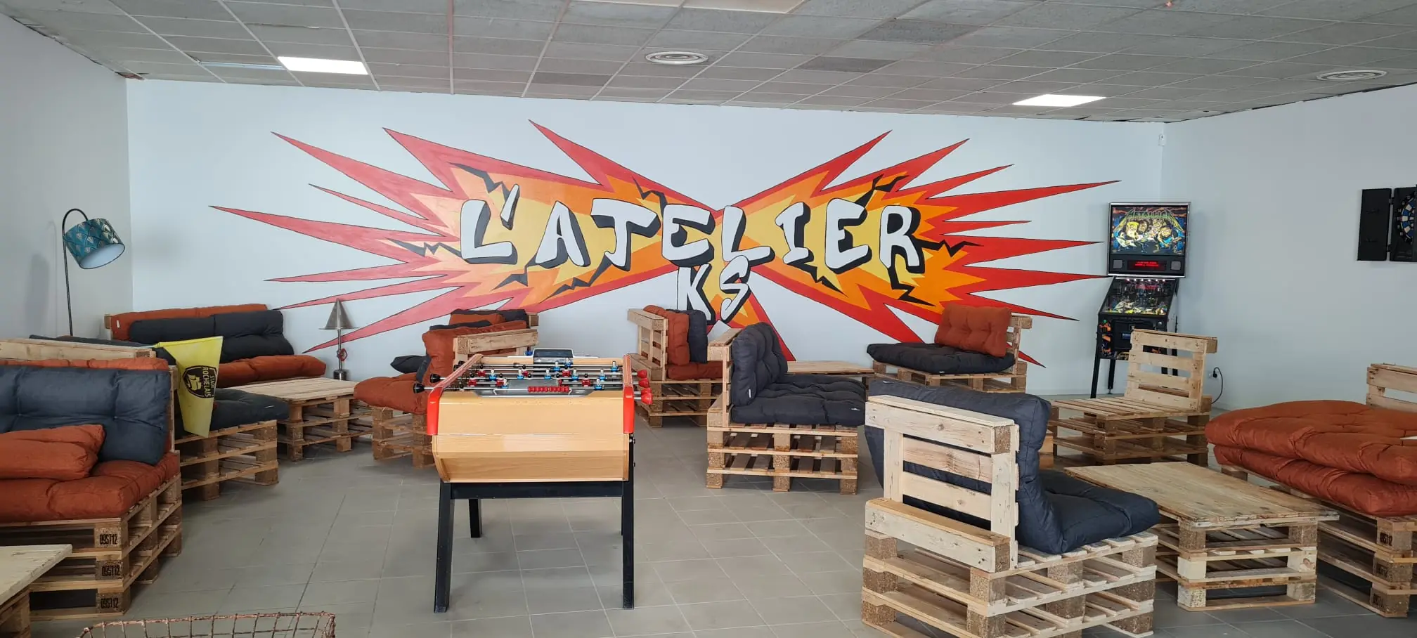 L'espace détente de l'AtelierKS à La Rochelle, vide : fauteuils et tables basses en palettes avec coussins orange et gris, un baby-foot, un flipper et une cible de fléchettes devant la fresque L'Atelier KS