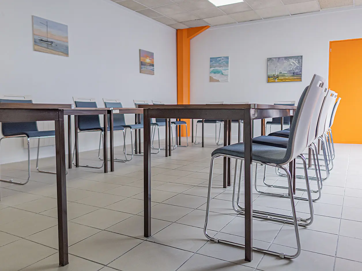 Tables et chaises de la salle de réunion de L'Atelier KS, quatre tableaux de bord de mer au mur, porte orange au fond.