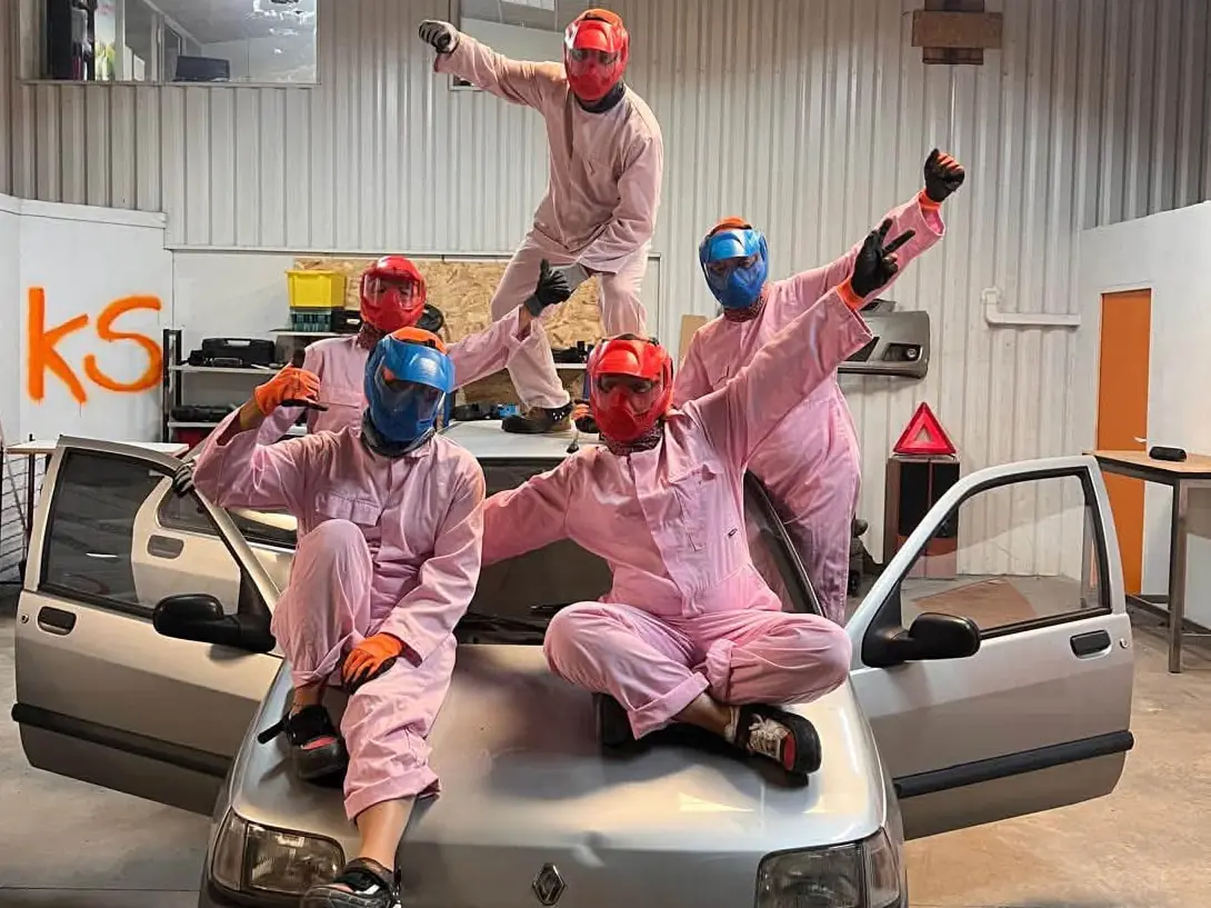 Cinq participants en combinaison rose, casque à visière et gants, posent sur une voiture avant la casse, dans le garage de L'Atelier KS à La Rochelle.